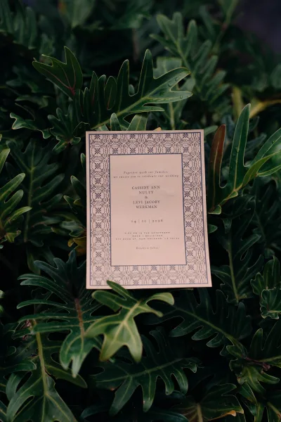 Wedding invitation with patterned border and navy lettering on a white card, styled on green foliage with tropical leaves background