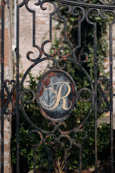 Wedding monogram sign with gold lettering on an oval plaque mounted to a wrought iron gate, framed by greenery and a brick wall