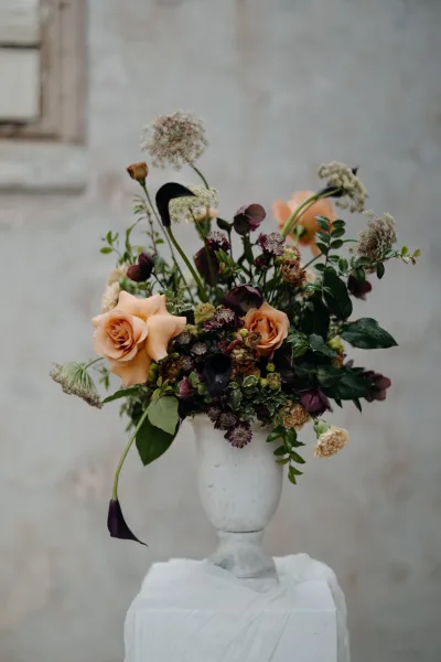 Wedding centerpiece in a ceramic urn vase with peach roses, dark purple blooms, and greenery on a white pedestal by a plaster wall window frame