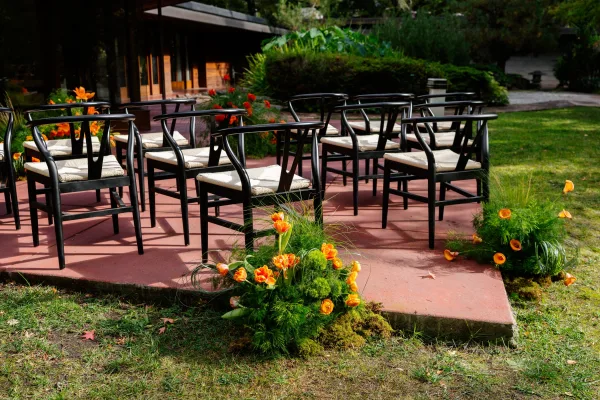 Ceremony seating with outdoor wedding ceremony chairs in black rows, woven seats, and orange ground florals with greenery on a garden patio lawn