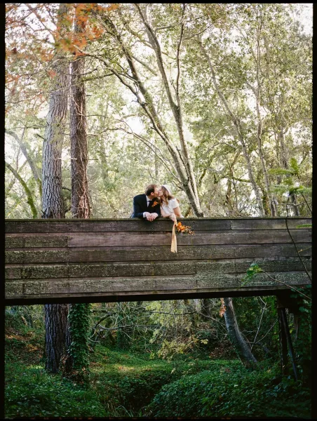 Wedding kiss as newlyweds kissing on a wooden bridge in the forest, bride holding a bouquet with long ribbon as they lean on railing