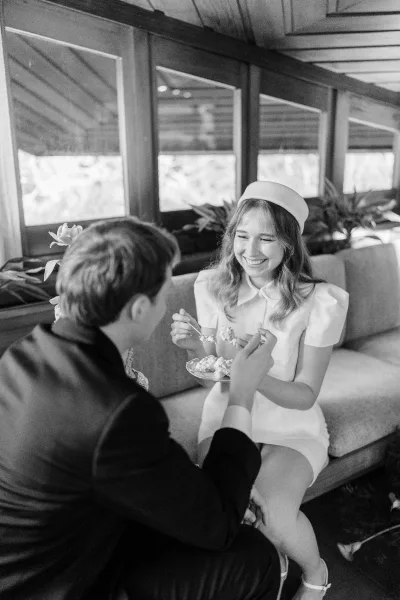 Couple portrait of newlyweds eating cake on a sofa, bride in a short white dress and pillbox hat in a window-lit lounge