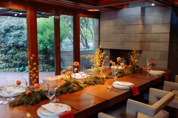 Reception tablescape with moss runner and orange floral centerpieces on a wood dining table, set near stone fireplace and garden-view doors