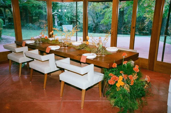 Reception tablescape with a long banquet table set on a wood dining table, greenery garland and bud vases by floor-to-ceiling windows overlooking a garden lawn