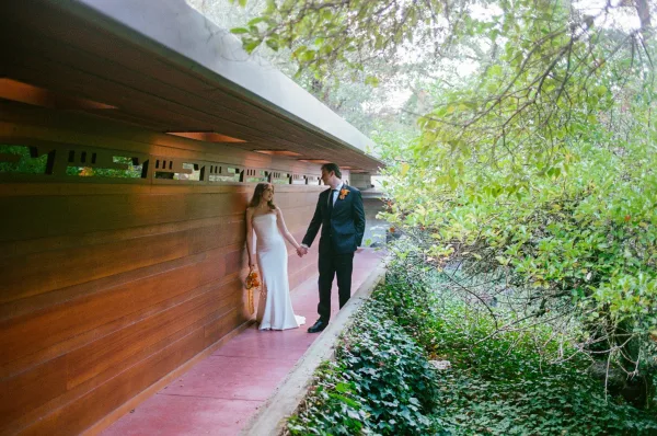 Couple portrait of bride and groom holding hands, bride with orange bouquet, walking by a wood wall along a leafy garden walkway