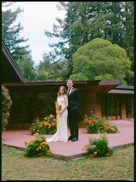 Couple portrait of bride in strapless dress holding an orange bouquet beside groom in black suit on patio by mid-century house and trees