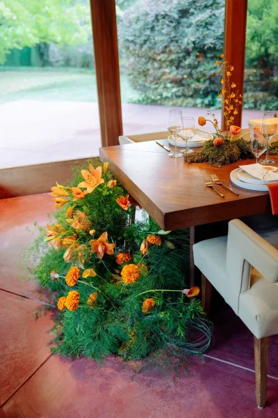 Reception tablescape with wedding table decor on a wood dining table, orange florals and moss runner by floor-to-ceiling windows facing garden lawn