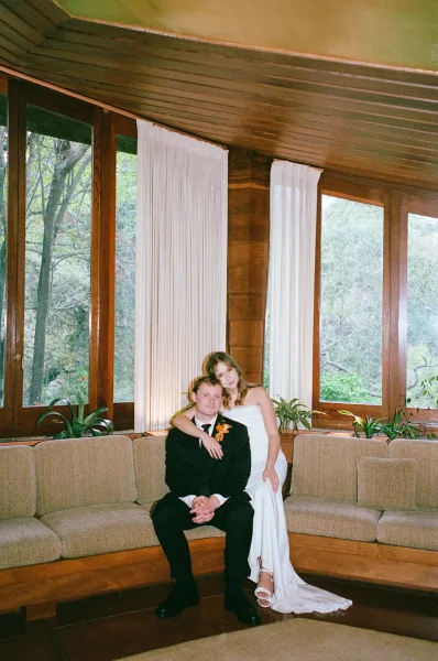 Couple portrait of bride sitting on groom’s lap on a sofa, strapless wedding dress and black suit in a wood-paneled room by windows