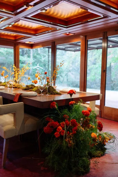 Reception tablescape with moss runner, orange blooms and bud vases on a wood table, set by floor-to-ceiling windows and trees