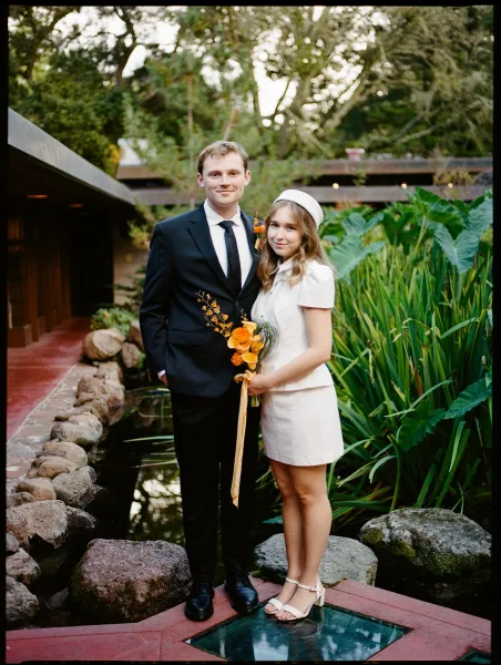Couple portrait of bride and groom standing by a garden pond, bride holding an orange bridal bouquet with ribbon, groom in black suit