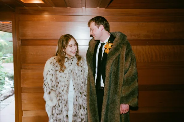 Couple portrait of bride and groom laughing in faux fur coats, gazing at each other in front of a wood paneled wall and window