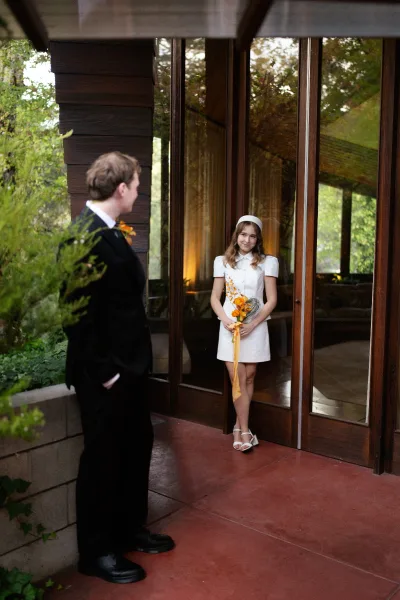 First look moment as groom sees bride in a short white mini wedding dress holding an orange bouquet by glass doors and greenery