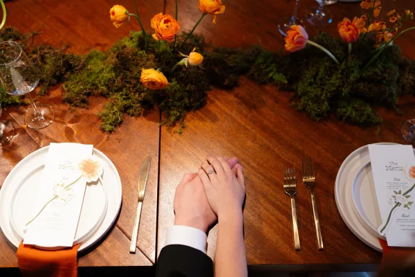 Reception tablescape with a moss table runner, orange flowers, gold flatware, printed menus on plates, and wine glasses on a wooden table
