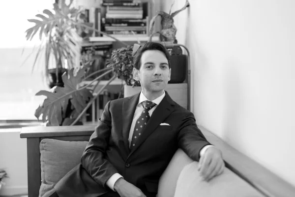 Groom portrait indoors in a suit with a neat pocket square, set against a bookshelf with houseplants and a window.