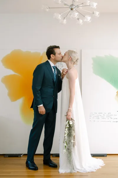 Wedding kiss portrait of a couple holding hands, the bride cradling a cascading bouquet, set against abstract art panels on a white wall.