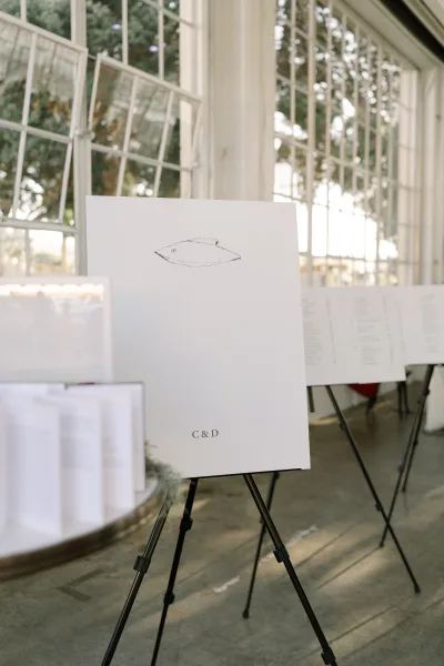 Wedding signage with a seating chart display on an easel, set indoors by expansive glass windows with trees beyond and a smooth concrete floor.