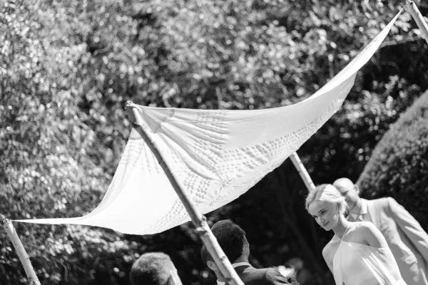Ceremony moment as the couple stands beneath a simple chuppah canopy during an outdoor garden ceremony among trees and hedges.