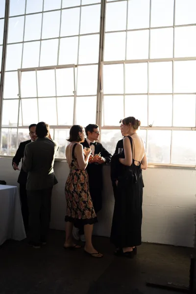 Wedding cocktail hour with guests mingling by industrial windows, a tuxedo among chic attire, city skyline visible beyond the glass.