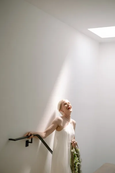 Bridal portrait of a laughing bride with a ribbon-wrapped bouquet, posed by a white staircase beneath a skylight