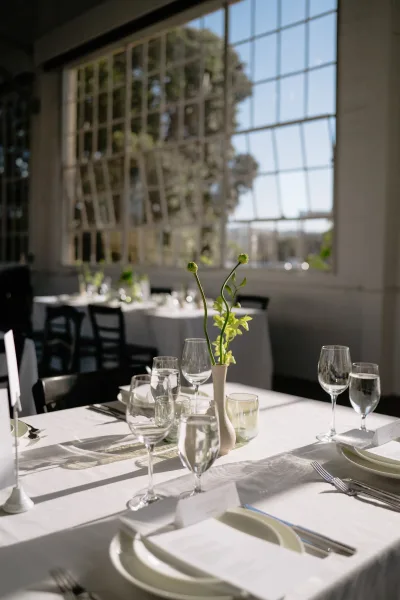 Reception tablescape for a minimalist wedding table with bud vase blooms, framed by large industrial windows and garden trees in daylight.
