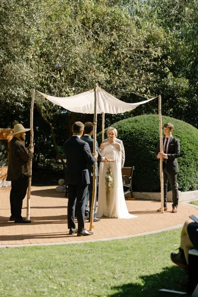 Ceremony moment at an outdoor garden ceremony as the couple stands beneath a wedding canopy, with garden trees and a brick path behind them.