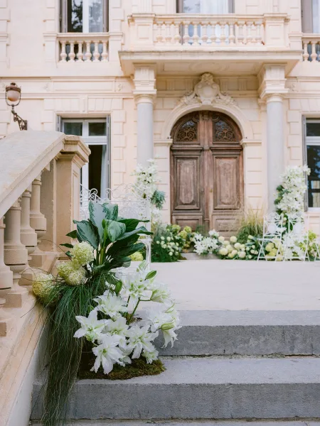 Outdoor ceremony decor with a staircase floral installation of white lilies flanking the stone staircase before a mansion facade with wooden doors.