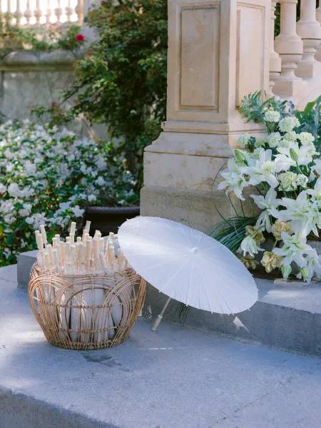 Wedding parasols arranged as a wedding parasol station in a wicker basket on stone steps beside a balustrade and garden bushes.