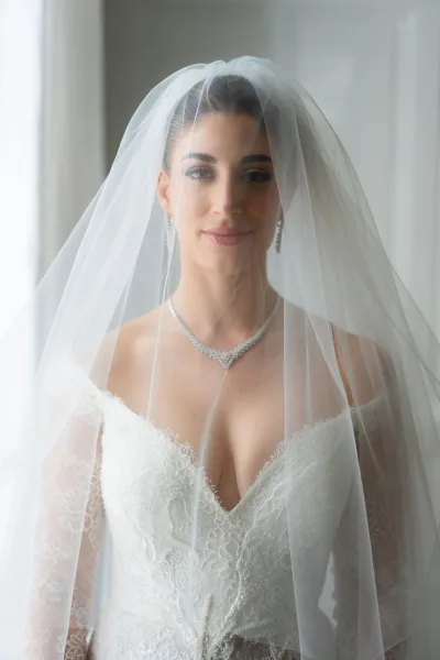 Bridal portrait of a bride under veil, gazing softly in window light, wearing a lace wedding dress near a white wall and sheer curtain.