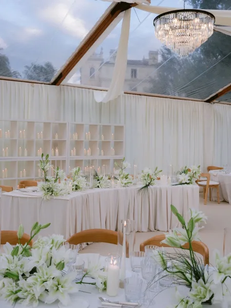 Reception tablescape with head table decor highlighted by white lilies beneath a clear-top tent with white drapery and sky visible overhead.