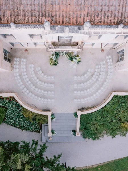 Ceremony setup for a courtyard ceremony layout with ghost chairs arranged on a stone terrace before a historic facade and balcony.