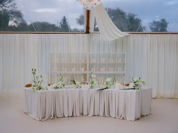 Reception tablescape with head table decor, white lilies amid low arrangements, under a clear tent ceiling as evening sky and trees softly glow.