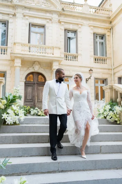 Couple portrait of bride and groom walking hand in hand on stone steps before a historic mansion facade, her veil trailing softly.