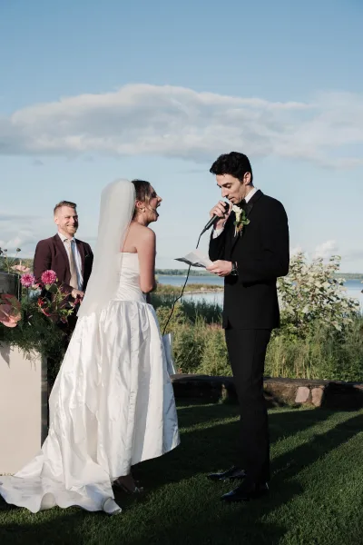 Wedding vows during an outdoor ceremony moment by a lakeside lawn, groom reading from vow paper as the bride laughs amid garden greenery.
