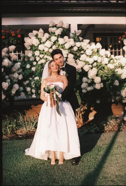Couple portrait of the bride and groom embracing, her veil trailing as they stand by blooming hydrangea bushes beside a house porch.