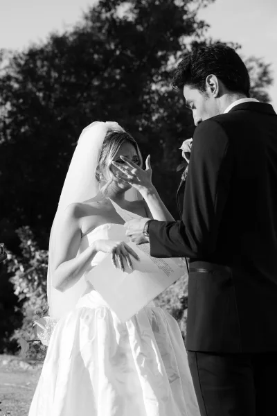 Vow exchange during an emotional ceremony moment, the bride holding vow letters while the groom reads, set amid garden trees under open sky.