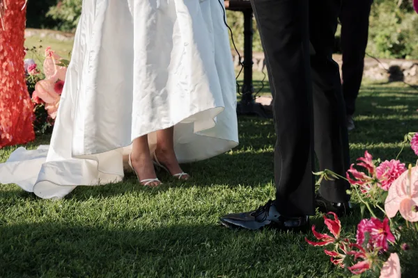 Ceremony moment showing a bride and groom shoes shot, with strappy bridal heels and patent leather groom shoes on grass lawn by garden greenery.