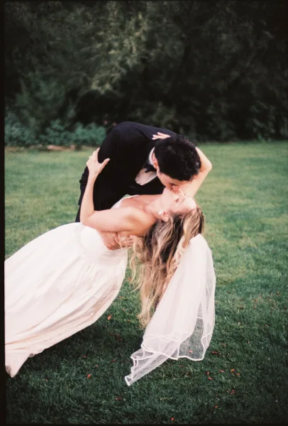 Wedding kiss portrait of a groom dipping his bride as her tulle veil flows on a grass lawn framed by garden trees and simple greenery.