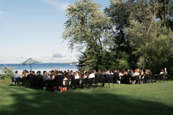 Outdoor ceremony setup shaded by an umbrella, overlooking a calm lake and tree line on a grass lawn for a serene lakeside wedding ceremony.
