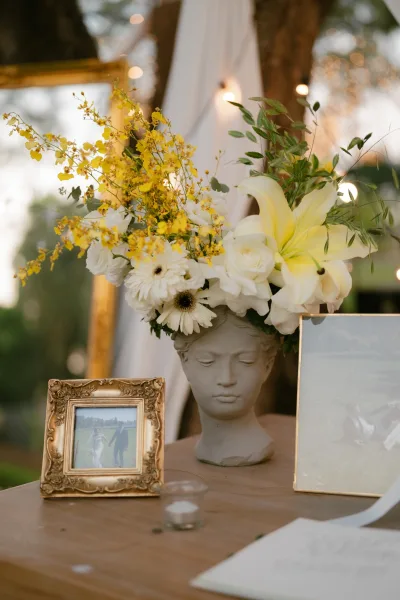 Wedding centerpiece featuring yellow orchids amid mixed blooms on a wooden table beneath garden trees, string lights, and white drapery.