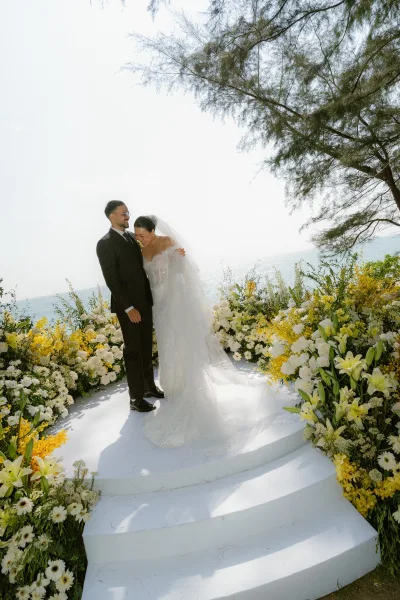 Couple portrait of the bride and groom laughing on a ceremony platform, with ocean and open sky beyond, tree branches framing the scene.