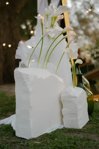 Floral arrangement of calla lily wedding flowers styled on sculptural white plinths amid garden trees and a grass lawn