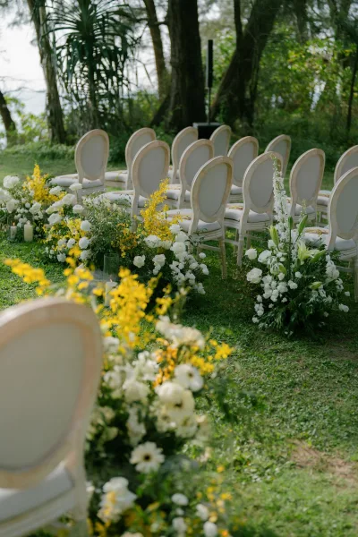 Ceremony aisle decor for an outdoor ceremony setup, featuring lush florals along the path on a grass lawn framed by tall trees.