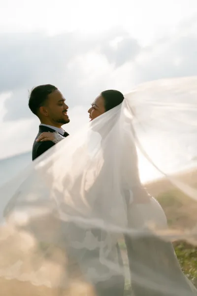Couple portrait of a bride and groom embrace under an airy bridal veil, backlit on a beach shoreline with soft ocean water and glowing sky.