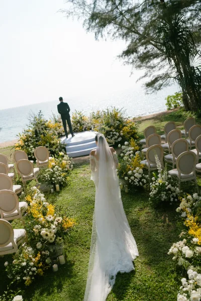 Ceremony processional with a bride walking down the aisle toward the groom on an oceanfront lawn, her long veil trailing as waves shimmer beyond.