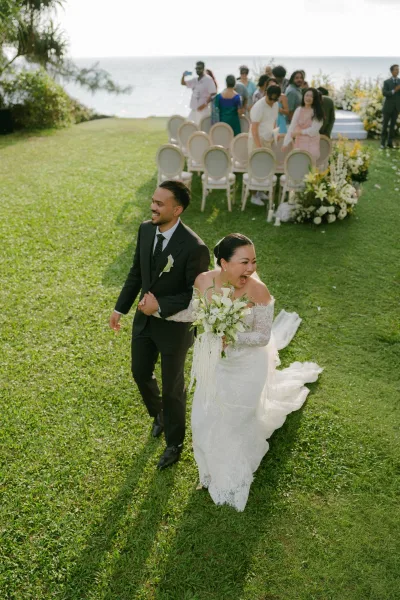 Wedding recessional as a just married exit, newlyweds walk the aisle hand in hand, her veil flowing, against an ocean and sky backdrop.