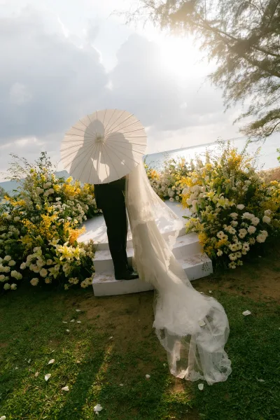 Couple portrait of a bride and groom silhouette under a parasol on a sunlit beach, the ocean shimmering behind and trees framing the horizon.