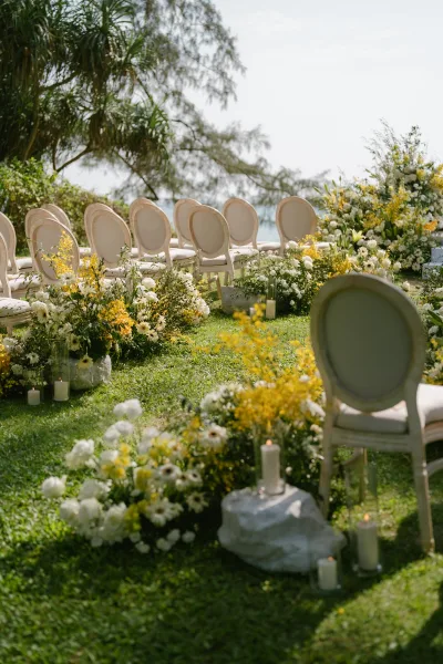 Ceremony setup showcasing outdoor ceremony decor with yellow and white florals lining a grass lawn aisle, framed by trees and the ocean.