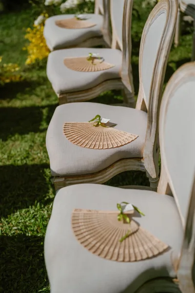 Ceremony seating with refined wedding chair decor, each seat holding a hand fan, on a grass lawn framed by garden greenery and yellow flowers.