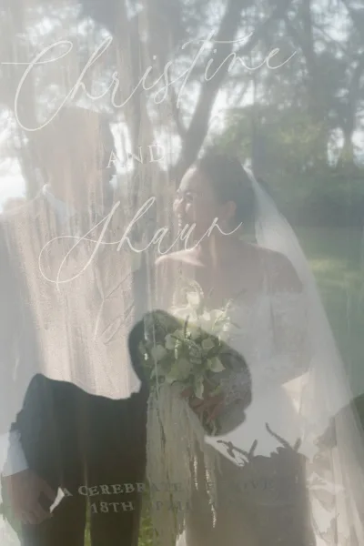 Couple portrait of bride and groom behind welcome sign, viewed through acrylic signage, smiling in a sunlit garden.