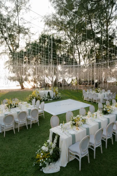 Outdoor reception decor beneath a string light canopy at a sunset beachfront lawn wedding, with elegant tables set among trees by the water.